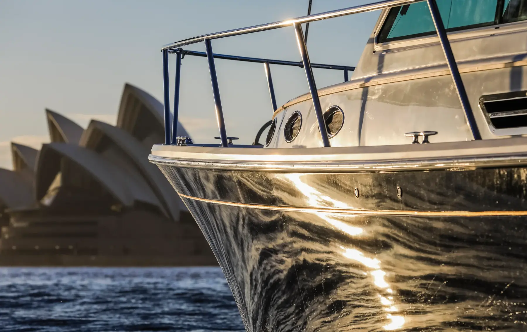 The bow of a Back Cove motor boat catching low golden light on Sydney Harbour, with the Opera House sails behind
