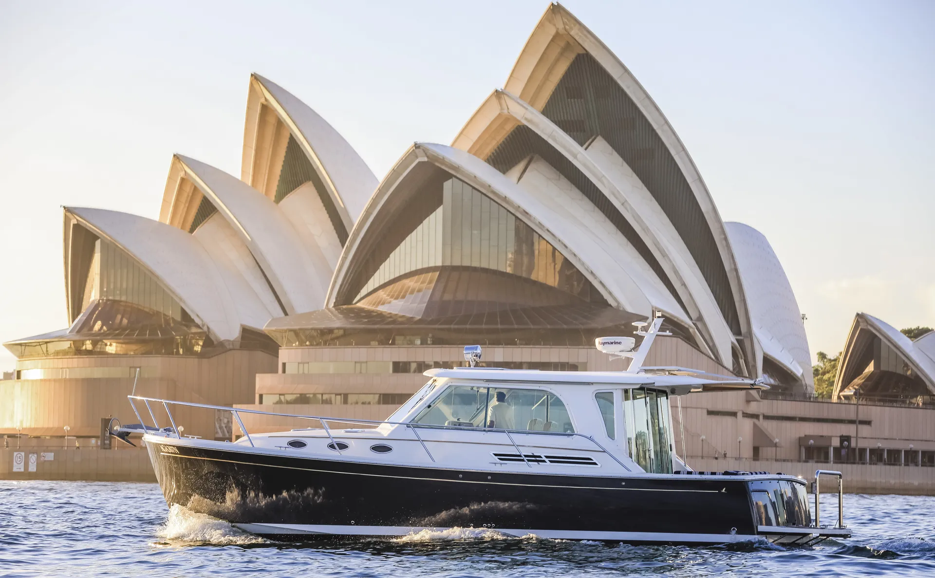 A navy-hulled motor yacht cruising past the Sydney Opera House at golden hour