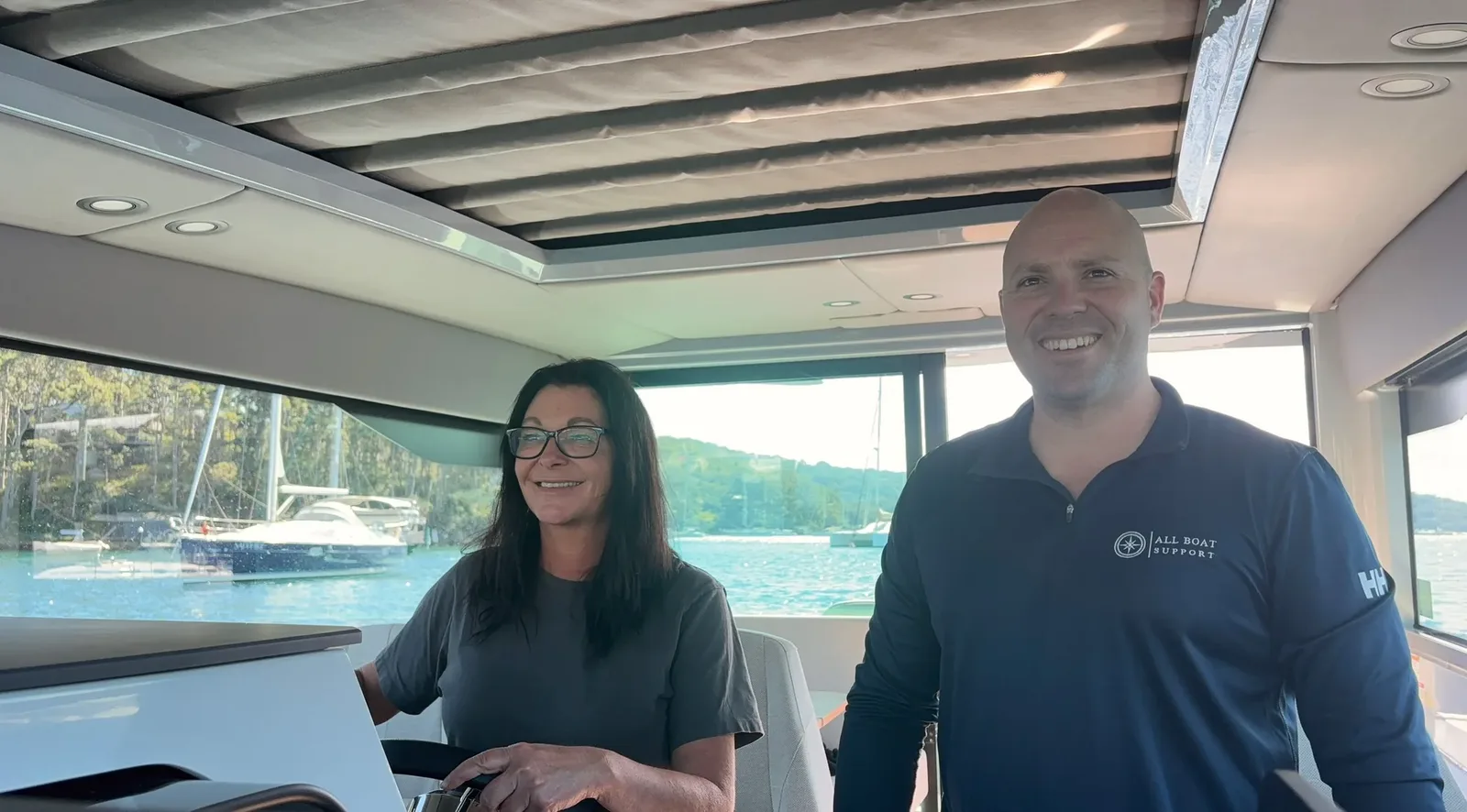 Two people at the helm inside a boat cabin, with moored sailboats and a forested hillside through the windows